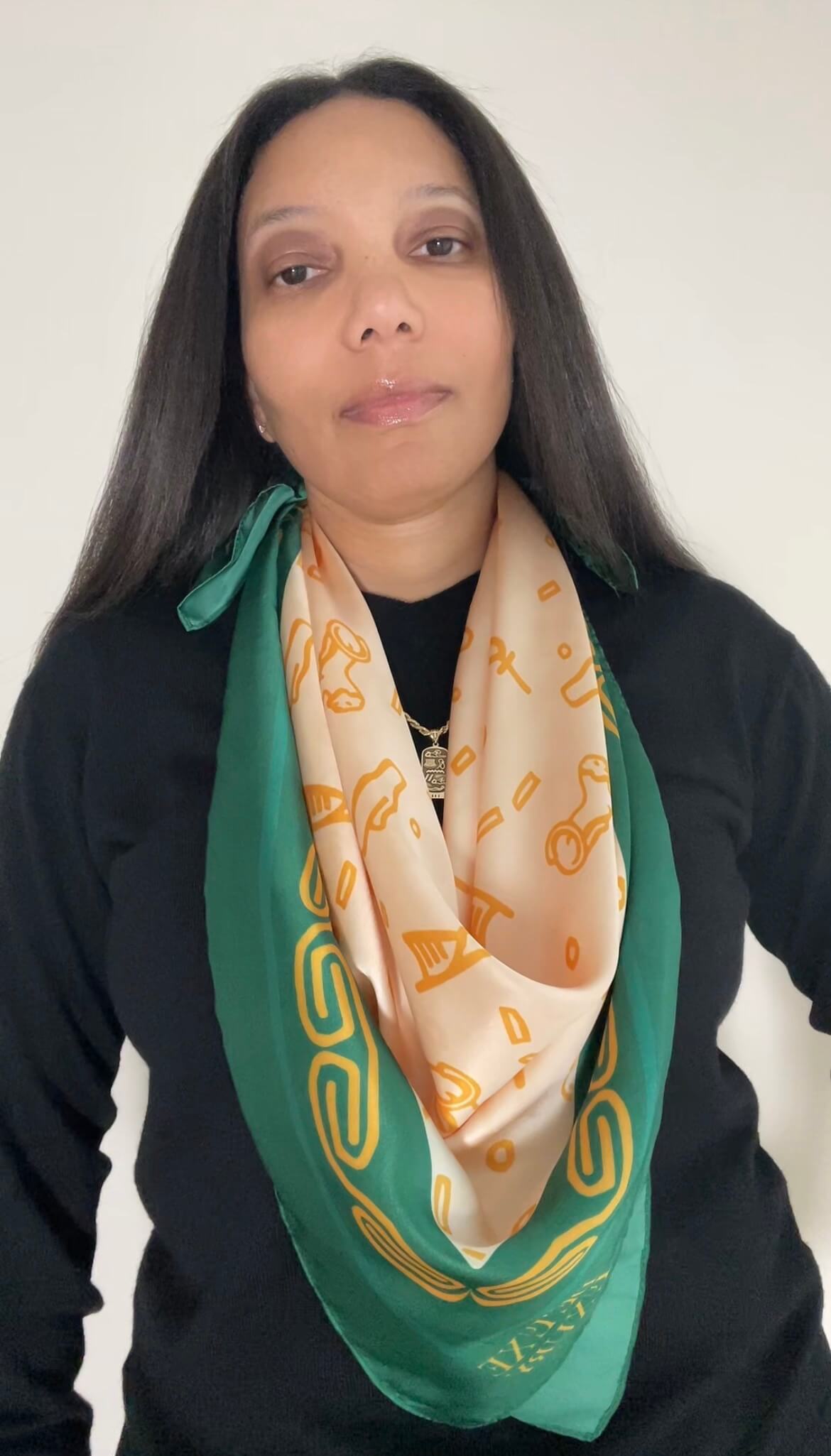 A woman with long black hair wearing a black long-sleeve top and the Origins Scarf in emerald, citrine and cream.