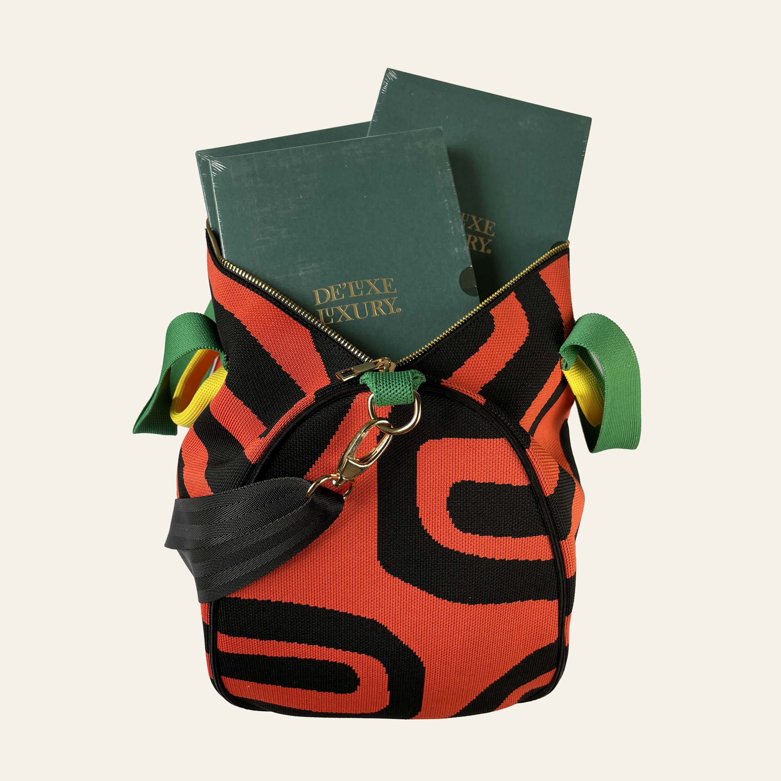 A small shoulder bag with a bold red and black abstract pattern, featuring green ribbon details on the sides and a black shoulder strap. The bag is shown open, filled with two dark green books, against a solid off-white background.