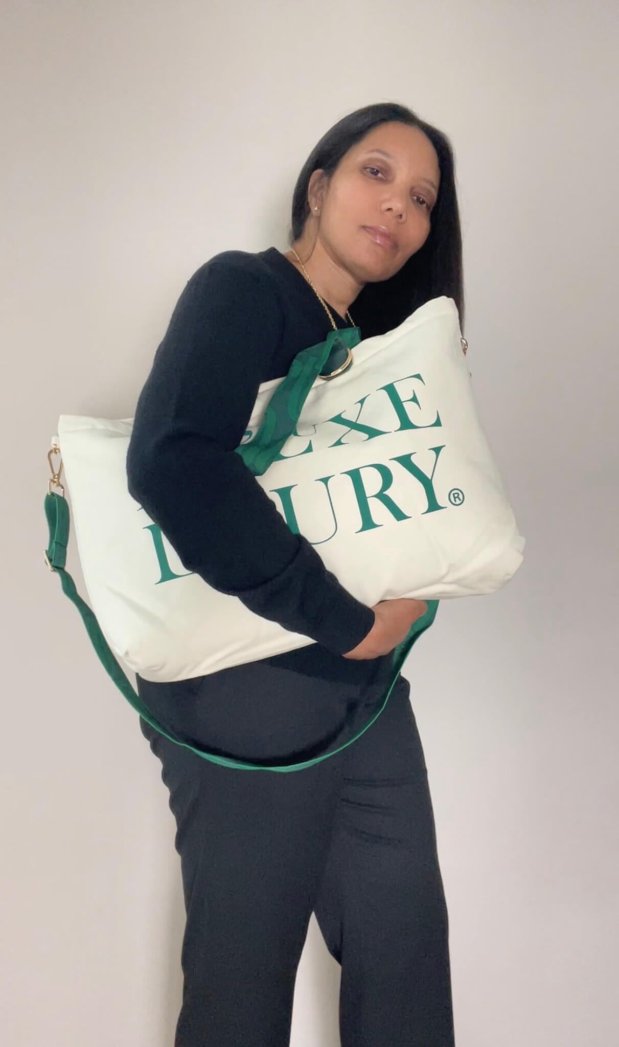 A woman wearing a black long-sleeve top and dark pants holds the Voyage tote with emerald text reading De'Luxe Luxury.